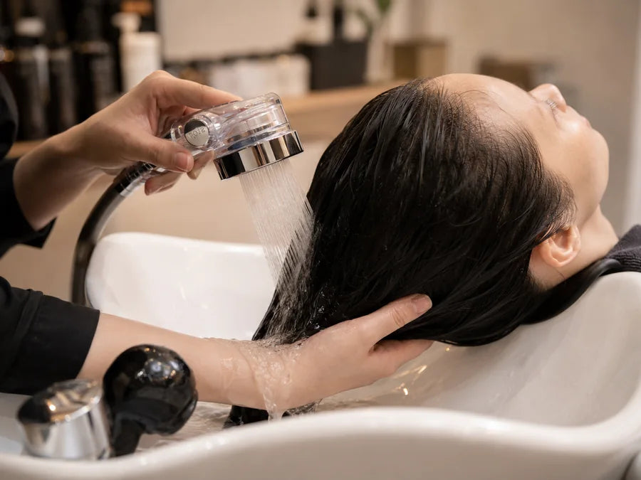 A salon stylist rinsing a client's hair with a handheld shower head.